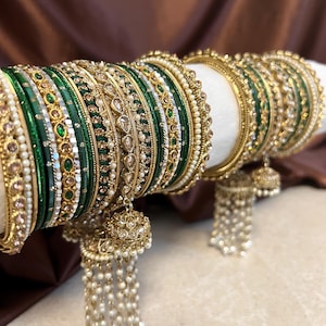 Può includere: Una collezione di braccialetti verdi e dorati, adornati con perle e pietre preziose. I braccialetti sono impilati, mostrando disegni intricati e dettagli dorati pendenti. I gioielli sono esposti su una superficie bianca contro uno sfondo marrone.