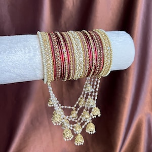 May include: A set of stacked bangles in shades of deep red, gold, and cream, featuring intricate metallic detailing and embedded clear stones. The set includes multiple thin bands and is accented with dangling gold-toned bell-shaped charms.