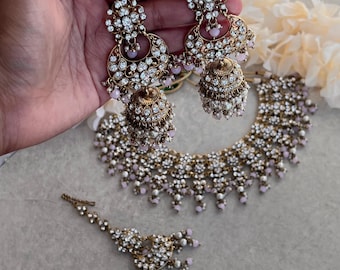 May include: A traditional Indian bridal jewelry set with a gold-tone necklace, earrings, and a maang tikka. The set features intricate designs with white stones and pink beads.