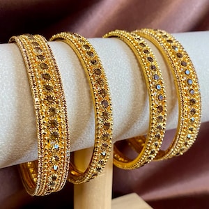 May include: Four gold-toned bangles featuring a central row of round, dark brown faceted stones. Each bangle is bordered by a delicate beaded texture on both sides, creating a consistent metallic finish across the set of circular bracelets.