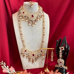 May include: A complete jewelry set featuring a necklace, long necklace, earrings, bracelet, and a headpiece. The gold-toned pieces are adorned with red and clear gemstones, displayed on a mannequin and a black hand.