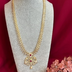 Puede incluir: Un collar dorado con un colgante detallado con piedras preciosas rojas. El collar se exhibe sobre un maniquí gris sobre un fondo rojo. El colgante tiene una piedra preciosa en forma de lágrima.