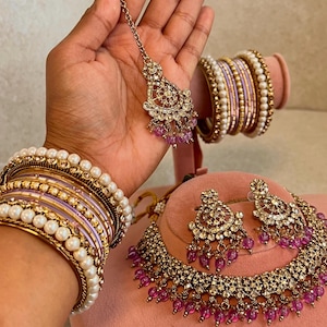 May include: An ornate gold-toned jewelry set featuring a necklace, earrings, and headpiece, adorned with clear crystals and small pink beads. The set is displayed with matching bangles, some with faux pearls, on a pink surface.
