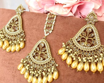 May include: A gold-tone bridal jewellery set with a teardrop-shaped pendant, earrings, and a maang tikka. The set features clear stones, white pearls, and a delicate floral design. The earrings and pendant have a fringe of cream-coloured pearls.