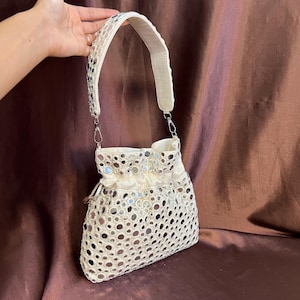 May include: Cream-coloured handbag with a shoulder strap. The bag is decorated with numerous small, round, reflective mirror accents. The strap and top of the bag have a textured design. The bag is held by a hand.