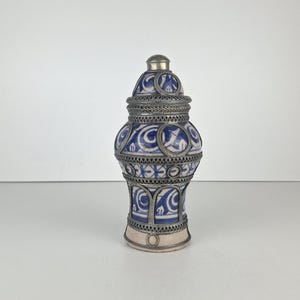 Könnte beinhalten: Ein Keramikgefäß mit Deckel, das ein blau-weißes Wirbelmuster aufweist. Das Gefäß ist von einem dekorativen, silberfarbenen Metallfiligran überzogen, das kreisförmige und bogenförmige Rahmen um die bemalte Keramikoberfläche bildet.