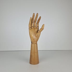 May include: A light brown wooden articulated hand model featuring movable fingers and a thumb, mounted on a cylindrical base. The natural wood grain is visible across the surface of the hand and the stand, showcasing a smooth, polished finish.