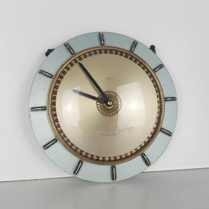 Puede incluir: Un reloj de pared redondo de color azul claro con detalles dorados y agujas negras. La esfera del reloj es blanca con un borde dorado y el texto "Made in Italy" está impreso en la esfera.