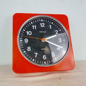 Può includere: Un orologio da parete quadrato rosso con un quadrante nero e numeri bianchi. L'orologio è realizzato da Kienzle e riporta la scritta "Quartz" sul quadrante.