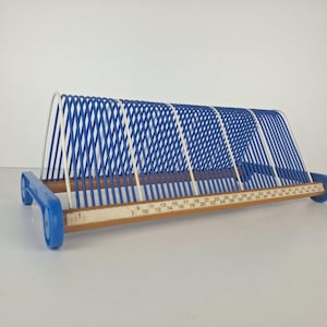 Può includere: Un portadischi in filo metallico blu e bianco con una base in legno dotata di una scala numerica stampata da 1 a 50. Il supporto ha un design a telaio triangolare con divisori in filo blu e tappi in plastica blu sui piedini della base.