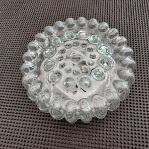 Pode incluir: Cinzeiro redondo de vidro transparente. O design apresenta uma série de protuberâncias arredondadas em forma de bolhas na borda e no centro. O vidro tem uma aparência texturizada, com pequenas bolhas.