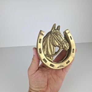 May include: A brass-colored metal horseshoe ornament featuring a raised relief of a horse head in the center. The horseshoe has a U-shape with visible nail holes along the outer edge and a detailed bridle on the horse head, creating a textured finish.
