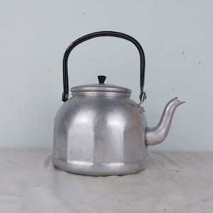 Puede incluir: Una tetera de aluminio plateado con asa negra y pomo de tapa negro. La tetera tiene un cuerpo redondeado con un pico ligeramente acampanado.