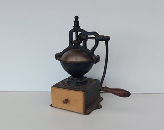Peut inclure: Moulin à café antique en fonte avec un tiroir en bois et une manivelle. Le moulin est noir et possède un bol en métal argenté.