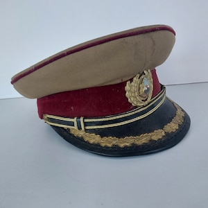 Può includere: Un cappello in stile militare color beige con una fascia rossa e bordi dorati. Il cappello ha un emblema dorato con una montagna e una stella.