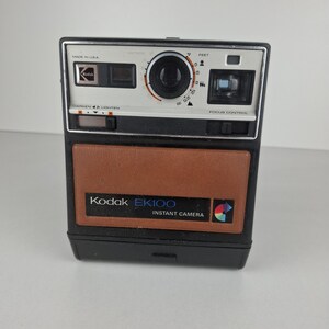 Puede incluir: Una cámara instantánea Kodak EK100 vintage. La cámara tiene un cuerpo negro con un panel frontal marrón. La sección superior es blanca y negra con una lente, visor y controles. El texto "Made in USA" es visible.