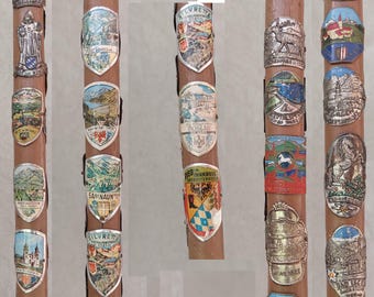Puede incluir: Un bastón de madera con múltiples insignias metálicas adjuntas. Las insignias están decoradas con varias imágenes y textos, que incluyen montañas, paisajes y paisajes urbanos.