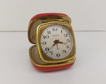 May include: A gold-colored alarm clock with a white face and black numbers. The clock is in a red case and says "EUROPA" on the face.