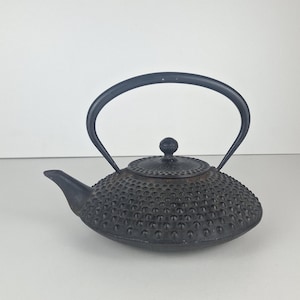 May include: A black cast iron teapot featuring a textured hobnail pattern across its rounded body, a matching lid with a small knob handle, a curved spout, and a high, arched metal handle that extends over the top of the vessel.