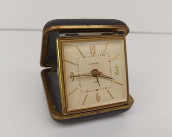 May include: A vintage Europa travel alarm clock in a black leatherette case. The clock face is square with gold-tone hands and numerals. The words "EUROPA" and "2 JEWELS" are printed on the face. The clock is open, revealing the face.