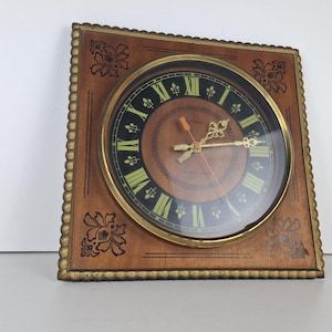 May include: Square wooden wall clock with a gold-tone bezel and a brown face. The clock features black Roman numerals, gold hands, and a decorative floral design on the wooden frame. The clock face is approximately 8 inches in diameter.