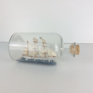 Puede incluir: Una botella de vidrio con un tapón de corcho que contiene un barco en miniatura blanco con velas azules y blancas. El barco flota en un mar azul y blanco.