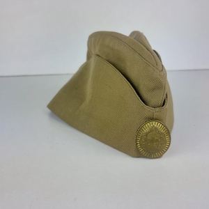 Può includere: Un cappello di stile militare color beige con un emblema circolare dorato sul lato.