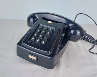 Może przedstawiać: Czarny telefon w stylu vintage z klawiaturą. Słuchawka jest połączona z podstawą za pomocą spiralnego przewodu. Telefon ma prostokątną podstawę i zakrzywioną słuchawkę. Klawiatura ma cyfry i symbole.