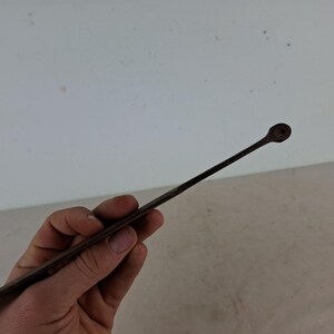 May include: A long, thin, metal tool with a rounded tip. The tool is held in a hand, and the background is a white wall.