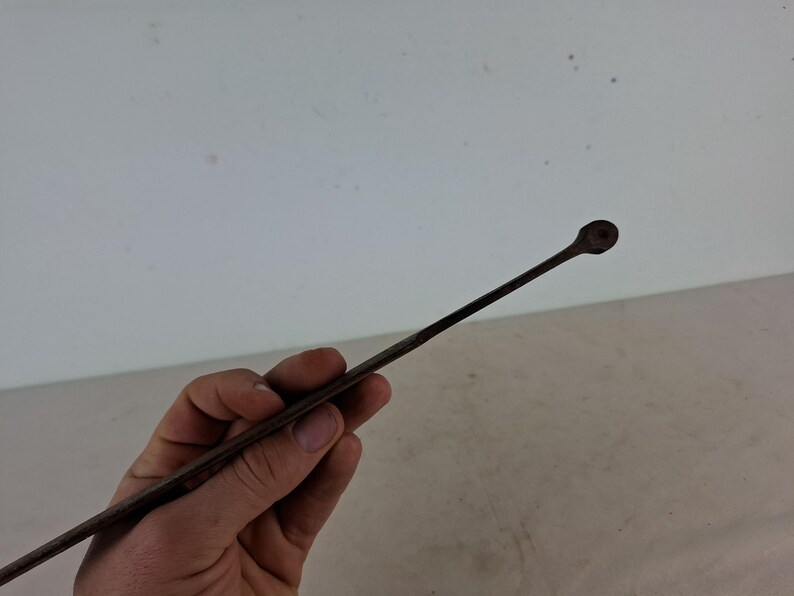 May include: A long, thin, metal tool with a rounded tip. The tool is held in a hand, and the background is a white wall.
