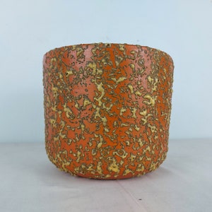 May include: A cylindrical orange planter with a textured surface and a speckled pattern of yellow and brown. The planter has a rounded base and is suitable for indoor or outdoor use.