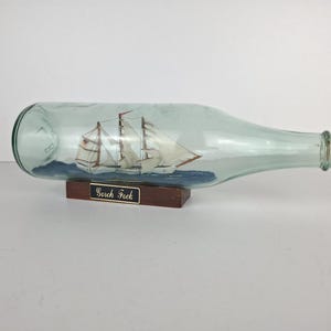Puede incluir: Una botella de vidrio transparente con tapón de corcho, que contiene una escena de velero en miniatura. El barco tiene velas blancas y está sobre un mar azul. La botella descansa sobre una base de madera con el texto "Serch Fock".