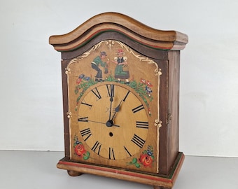 May include: A wooden clock with a painted face and Roman numerals. The clock has a painted scene of two people in traditional clothing. The clock is on a white background.