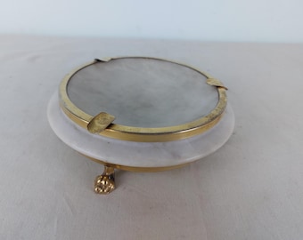 May include: A white marble ashtray with a gold metal rim and three gold feet. The ashtray has a smooth, polished surface.