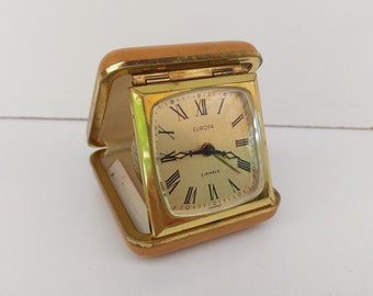 May include: A gold-colored alarm clock with a square face and a hinged lid. The clock face has black Roman numeral hour markers, black hands, and the words "EUROPA" and "2 JEWELS GERMANY".