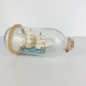 Puede incluir: Una botella de vidrio con un tapón de corcho y un pequeño modelo de barco en su interior. La botella está envuelta con una cuerda marrón.