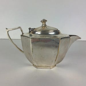 Puede incluir: Una tetera de plata con tapa abatible y cuerpo octogonal. La tetera tiene un pico y un asa.