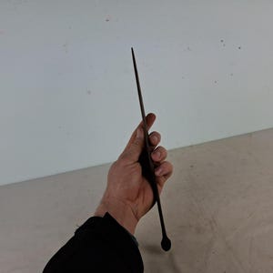 May include: A long, thin, black metal rod with a rounded tip. The rod is held in a person's hand.
