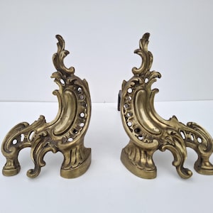 May include: A pair of antique brass and gold-toned fireplace andirons. Each andiron has an ornate, curved design with scrollwork and decorative cutouts. The andirons have a sturdy base and are designed to hold logs.