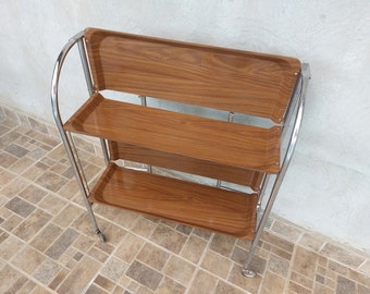May include: A three-tiered serving cart with a chrome frame and brown wood-grain trays. The cart has a curved handle and small wheels for easy mobility. The trays are rectangular and have a raised edge.