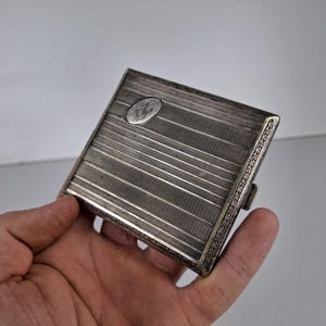 Könnte beinhalten: Ein silbernes Zigarettenetui mit einem dekorativen Rand und einer erhabenen ovalen Form auf der Vorderseite. Das Etui hat eine strukturierte Oberfläche und ein Scharnier an der Seite.