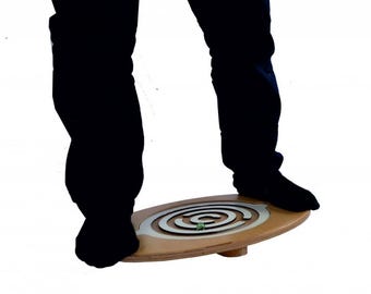 May include: An oval-shaped wooden balance board featuring a silver-colored maze pattern on its surface. A small green marble is positioned within the maze tracks. The board is designed for standing, with a central pivot point underneath.