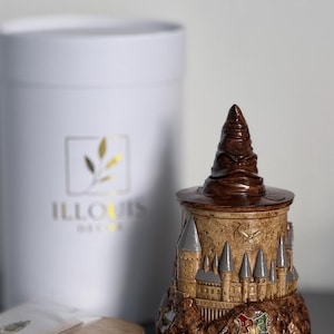 May include: A decorative ceramic container shaped like Hogwarts Castle and the Sorting Hat. The container is brown, tan, and gray with the Hogwarts crest and a dark brown Sorting Hat lid. A white cylindrical box with the logo "ILLOUIS DECOR" is in the background.