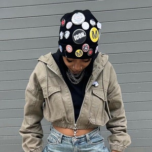 May include: A person wearing a black beanie with various pins and patches, a green cropped jacket, and blue jeans.