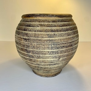 May include: A large, round, ceramic vase with a textured, horizontal striped pattern. The vase is primarily brown with lighter beige stripes and a dark brown rim. The vase has a wide body and a slightly narrower base.
