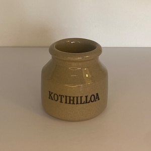 Pode incluir: Um pequeno pote de cerâmica bege com uma abertura larga e um corpo arredondado. A palavra "KOTIHILLOA" está impressa em preto na frente. O pote é provavelmente usado para armazenar alimentos.