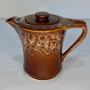 Peut inclure: Théière en céramique marron avec couvercle et anse. La théière présente un motif tacheté et texturé avec un glaçage brun foncé. Le bec verseur est sur le côté gauche et la anse est incurvée.