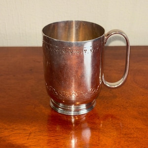 May include: A silver-coloured mug with a handle, featuring decorative floral patterns near the top and bottom. The mug sits on a small, circular base. The mug is made of a shiny metal.
