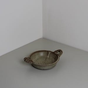 May include: A small, round, ceramic bowl with two decorative handles. The bowl is a muted green-brown colour with a textured surface and a spiral pattern in the centre. The handles have a woven rope design.