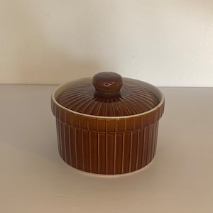 Op de afbeelding: Een bruine keramische ramekin met een deksel. De ramekin heeft verticale geribbelde zijkanten en een ronde, geribbelde deksel met een kleine knop. De binnenrand is lichter van kleur. De ramekin is ontworpen voor het bakken en serveren van individuele porties.